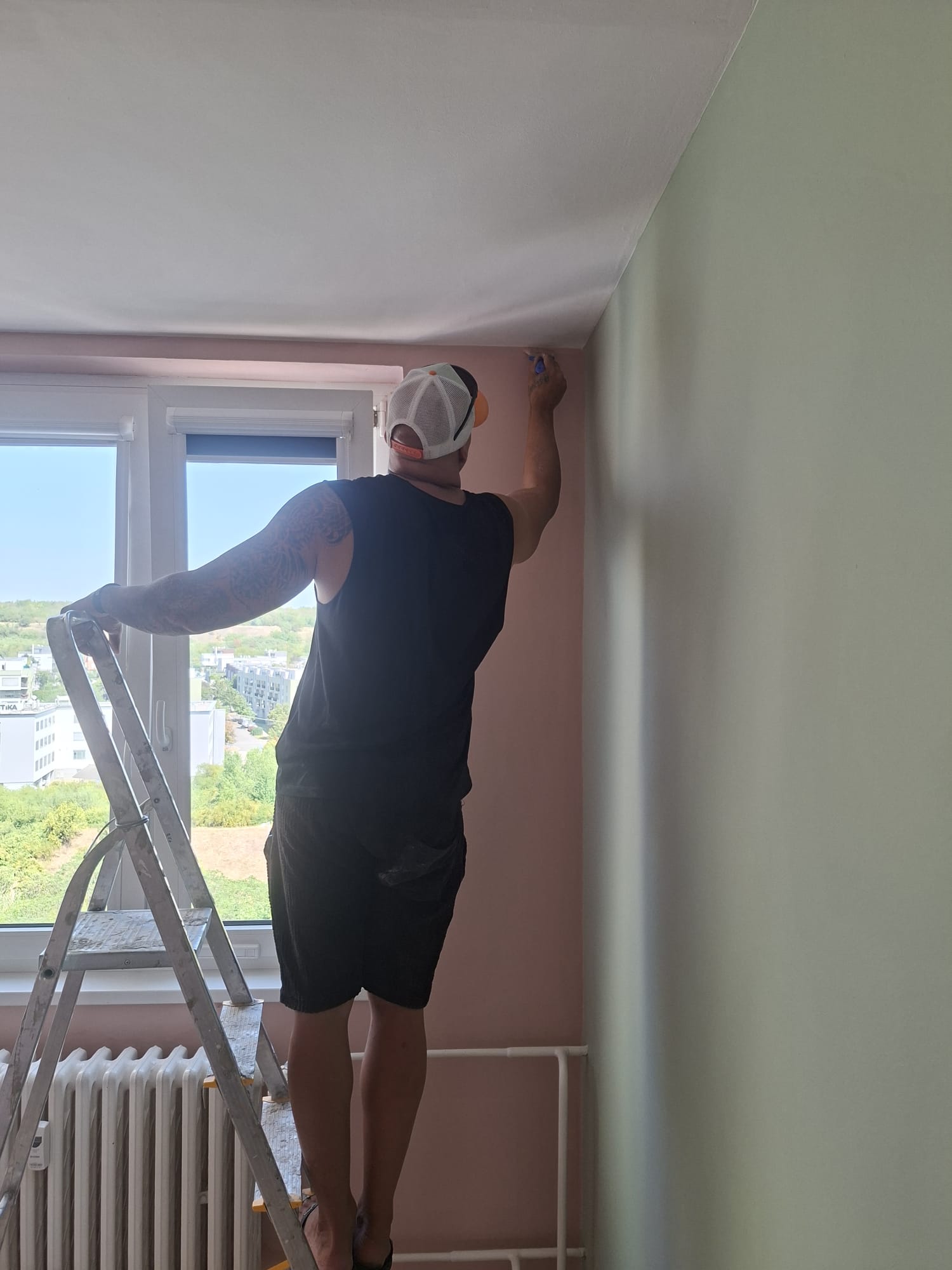 Malíř natírá hranu stropu v pokoji s růžovou a zelenou stěnou